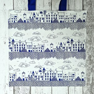 Puede incluir: Bolso tote blanco con un dise&ntilde;o de paisaje urbano ilustrado en azul y blanco. El dise&ntilde;o presenta edificios, &aacute;rboles, nubes y un patr&oacute;n similar a una valla. El bolso tiene asas azules.