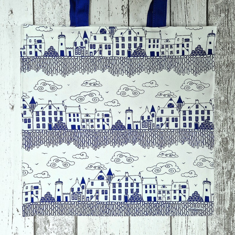 Puede incluir: Bolso tote blanco con un dise&ntilde;o de paisaje urbano ilustrado en azul y blanco. El dise&ntilde;o presenta edificios, &aacute;rboles, nubes y un patr&oacute;n similar a una valla. El bolso tiene asas azules.