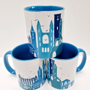 May include: Three blue and white ceramic mugs with a cityscape design. The mugs have blue rims and handles. The cityscape design features buildings, a clock tower, and birds.