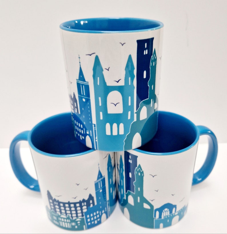 May include: Three blue and white ceramic mugs with a cityscape design. The mugs have blue rims and handles. The cityscape design features buildings, a clock tower, and birds.