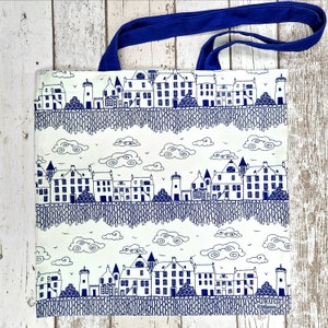Puede incluir: Bolso tote blanco con un dise&ntilde;o de paisaje urbano azul. El bolso presenta un patr&oacute;n repetitivo de edificios, nubes y un asa azul. Los edificios est&aacute;n detallados con ventanas y puertas, y las nubes est&aacute;n estilizadas.