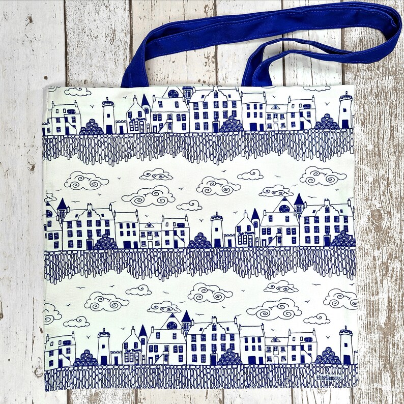 Puede incluir: Bolso tote blanco con un dise&ntilde;o de paisaje urbano azul. El bolso presenta un patr&oacute;n repetitivo de edificios, nubes y un asa azul. Los edificios est&aacute;n detallados con ventanas y puertas, y las nubes est&aacute;n estilizadas.