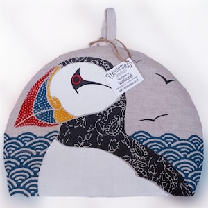 May include: A white linen tea cosy with a colourful puffin design. The puffin is facing the right side of the image. The cosy has a loop for hanging. The text "Thistlemoss Originals Handmade in Scotland www.thistlemoss.co.uk" is on a tag attached to the cosy.
