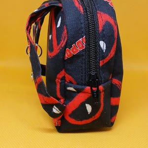 Puede incluir: Bolsa negra con cremallera con un patr&oacute;n de Deadpool rojo y blanco. La bolsa tiene una correa negra con un anillo plateado.