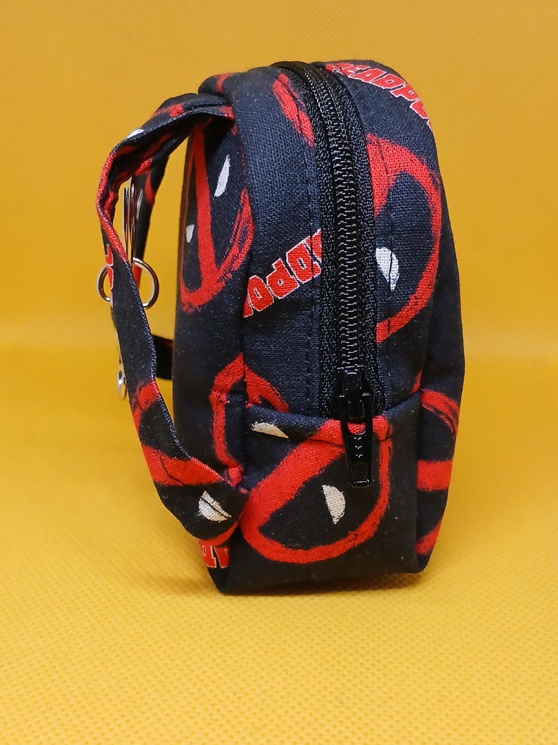 Puede incluir: Bolsa negra con cremallera con un patr&oacute;n de Deadpool rojo y blanco. La bolsa tiene una correa negra con un anillo plateado.