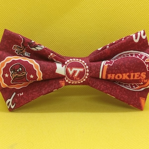May include: A red bow tie featuring a Virginia Tech Hokies logo and mascot. The bow tie is made of fabric and has a white background with orange and red accents.
