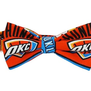 May include: A vibrant orange bow tie featuring the OKC Thunder logo in white, blue, and yellow. The bow tie has a repeating pattern of the team name and logo. The background is white.
