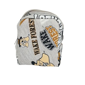 May include: A small, gray backpack with a repeating pattern of the words "WAKE FOREST" and a cartoon character in black, gold, and white. The backpack has a zipper closure and a small handle.