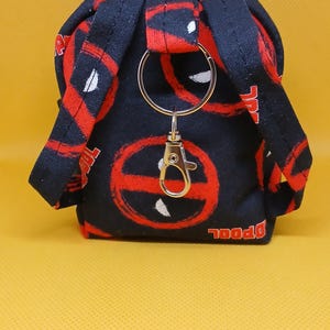 May include: A small black and red backpack keychain with a silver metal ring and clasp. The backpack has a Deadpool logo pattern.