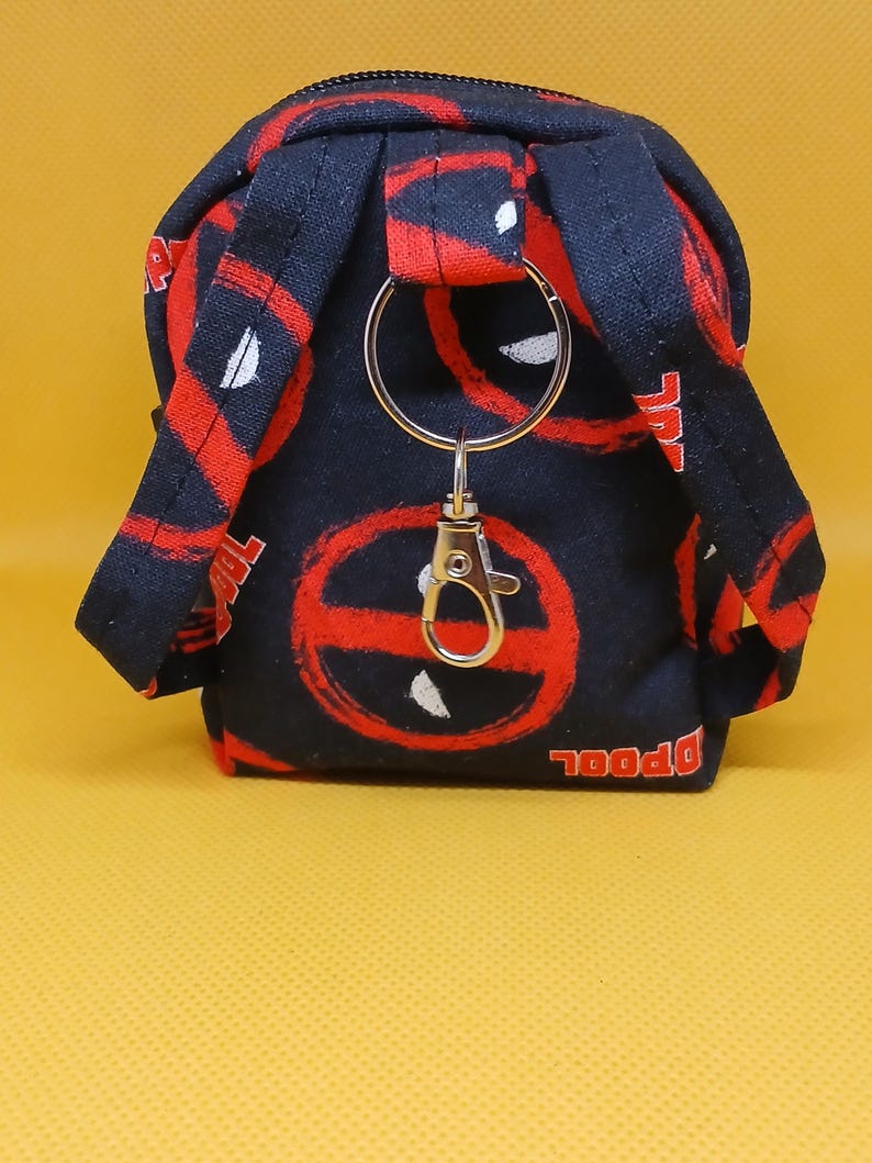 Puede incluir: Un peque&ntilde;o llavero de mochila negro y rojo con un anillo y un cierre de metal plateado. La mochila tiene un patr&oacute;n de logotipo de Deadpool.