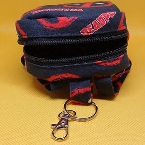Puede incluir: Una peque&ntilde;a mochila negra y roja con cierre de cremallera y un llavero plateado. La mochila tiene un patr&oacute;n repetido de la palabra "DEADPOOL" y un dise&ntilde;o de calavera roja y negra.