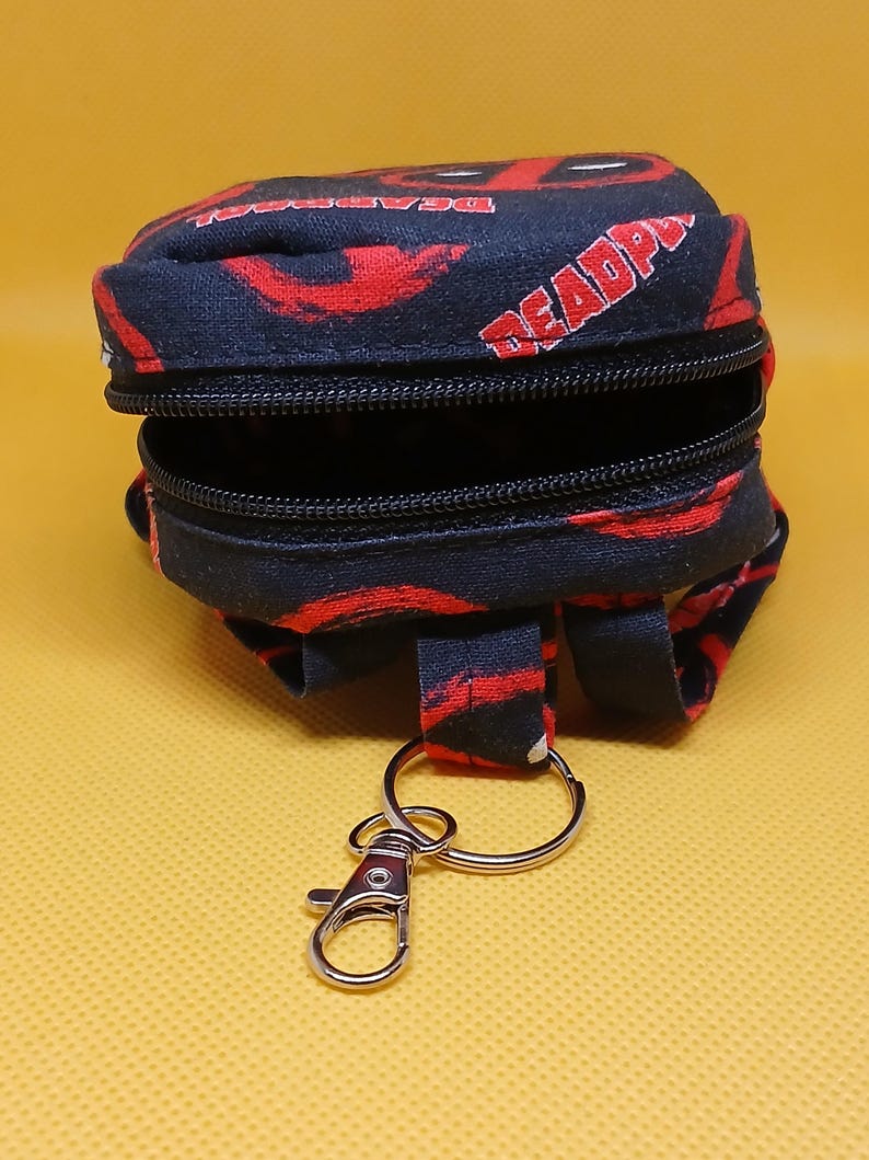 Puede incluir: Una peque&ntilde;a mochila negra y roja con cierre de cremallera y un llavero plateado. La mochila tiene un patr&oacute;n repetido de la palabra "DEADPOOL" y un dise&ntilde;o de calavera roja y negra.