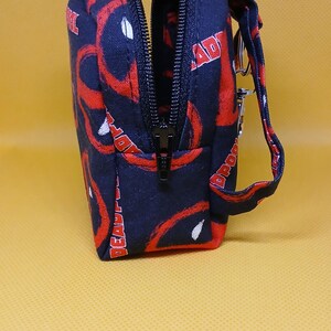 Puede incluir: Una bolsa negra con cremallera con un estampado de Deadpool rojo y blanco. La bolsa tiene una correa negra con un llavero plateado.