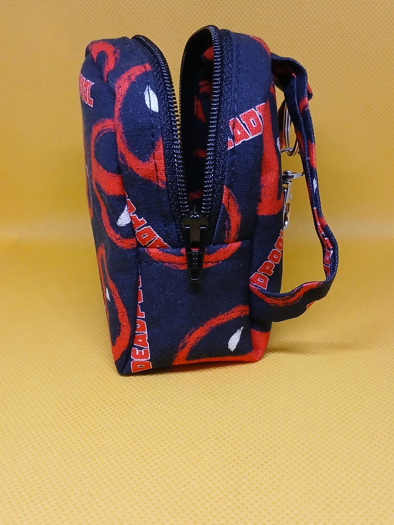 Puede incluir: Una bolsa negra con cremallera con un estampado de Deadpool rojo y blanco. La bolsa tiene una correa negra con un llavero plateado.