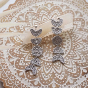 Puede incluir: Un par de pendientes de ganchillo plateados con un diseño de luna creciente. Los pendientes cuelgan de una superficie de madera con un patrón de mandala.