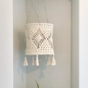 Op de afbeelding: Een witte gehaakte lampenkap met een geometrisch patroon en kwastjes die aan een wit plafond hangen. Een groene varenplant in een geweven mand staat op een geweven dienblad met dennenappels.