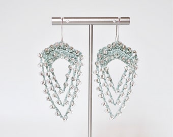 May include: A pair of silver and light blue beaded earrings hanging from a stand. The earrings are made of multiple layers of beads and have a teardrop shape.