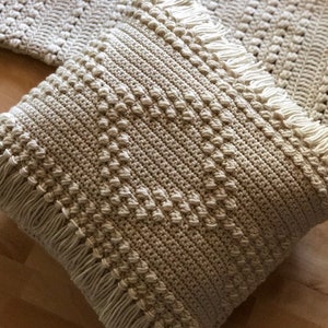 May include: A cream-colored crocheted pillow with a fringe border. The pillow has a textured pattern and is made with a thick yarn.