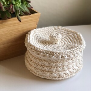 May include: A white crocheted storage basket with a lid. The basket has a round shape and a textured design. The lid has a small knob on top.