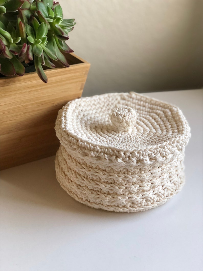 May include: A white crocheted storage basket with a lid. The basket has a round shape and a textured design. The lid has a small knob on top.