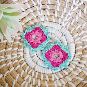 May include: A pair of crocheted earrings with a pink and turquoise color scheme. The earrings are square shaped with a flower design in the center.