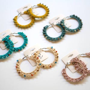 May include: A collection of handmade hoop earrings in various colors, including teal, yellow, beige, and pink. Each pair features a woven design with small, colorful beads. The earrings are displayed on white cards.