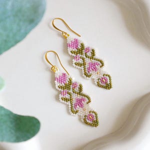 May include: A pair of beaded earrings with a floral design. The earrings have a white background with pink, green, and gold accents. The earrings are suspended from gold-coloured hooks.