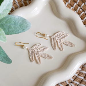 Puede incluir: Un par de pendientes de hojas de ganchillo beige con ganchos dorados y una pequeña perla blanca. Los pendientes se exhiben en un plato de cerámica blanco con bordes ondulados, con un fondo tejido.