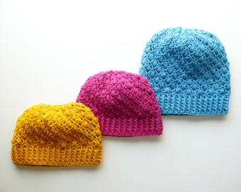 May include: Three handmade crochet beanies in vibrant colors: yellow, fuchsia, and turquoise. Each hat features a textured, raised stitch pattern and a ribbed brim. The beanies are displayed on a white background, showcasing their individual colors and textures.