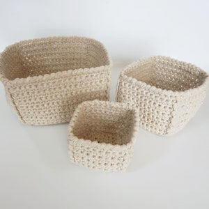 May include: Three crocheted baskets in various sizes, all in a cream colour. The baskets are made of a thick, textured yarn and have a square shape.