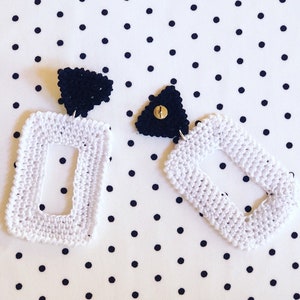 May include: A pair of white crocheted earrings with a black triangle accent. The earrings are shaped like a rectangle with a smaller rectangle cut out in the center.