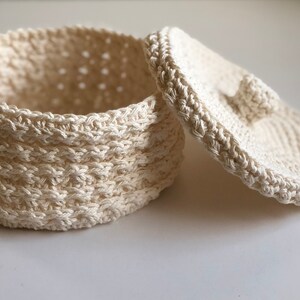 May include: A white crocheted storage basket with a lid. The basket is round and has a textured design. The lid has a small knob on top.