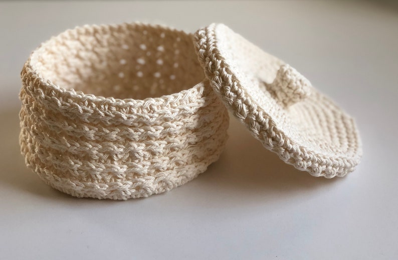 May include: A white crocheted storage basket with a lid. The basket is round and has a textured design. The lid has a small knob on top.