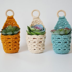 May include: Three crocheted hanging planters with wooden rings. The planters are in mustard yellow, cream, and teal. Each planter has a small succulent plant inside.