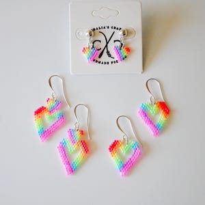 May include: Rainbow heart-shaped earrings with silver hooks. The earrings are made of small, colorful beads arranged in a heart design. A pair of stud earrings with the same design is displayed on a white card with text.