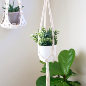 Può includere: Un portavasi in macramé bianco con un vaso bianco e una pianta verde appesa a un anello di legno. Il portavasi ha un lungo fiocco in basso.