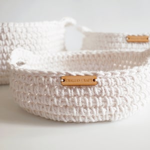 Puede incluir: Tres cestas blancas de ganchillo de diferentes tamaños. La cesta delantera tiene asas y una etiqueta de madera con "CHALLAS CRAFT". Las cestas están hechas de hilo grueso y texturizado, adecuadas para guardar o decorar.
