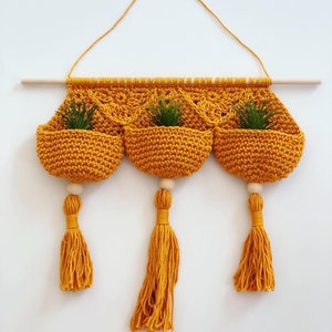 May include: A macrame plant hanger with three yellow crocheted pockets and wooden beads. The hanger has three tassels and is hanging from a wooden dowel.