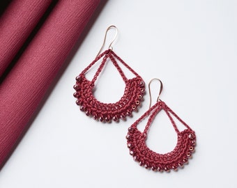 May include: A pair of maroon crocheted earrings with small beads hanging from the bottom. The earrings are in the shape of a teardrop with a curved top.