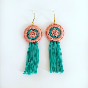 May include: A pair of crocheted earrings in a peach and teal colour scheme. The earrings feature a round, crocheted design with a tassel.