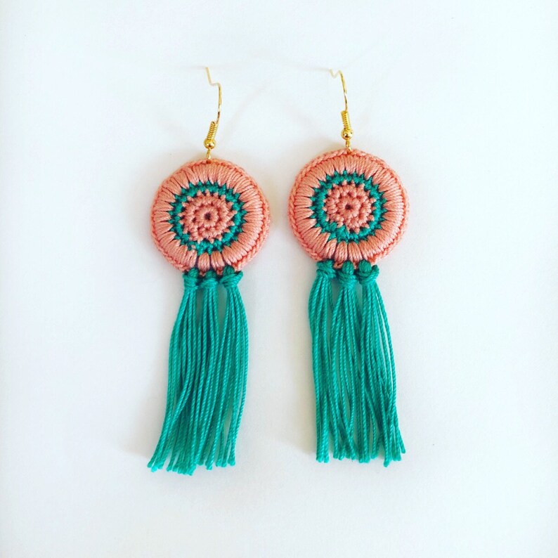 May include: A pair of crocheted earrings in a peach and teal colour scheme. The earrings feature a round, crocheted design with a tassel.