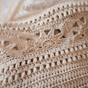 May include: Close-up of a beige crocheted throw blanket with a delicate, intricate pattern. The blanket has a soft, textured surface and is made with a fine yarn.