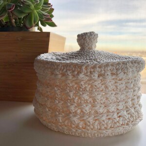 May include: A white crocheted basket with a lid. The basket is made with a textured stitch pattern and has a round shape.