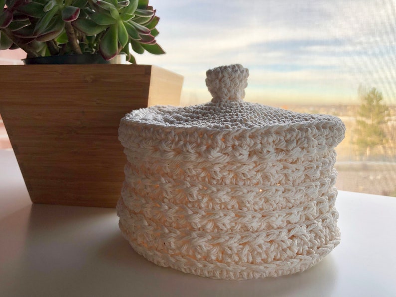 May include: A white crocheted basket with a lid. The basket is made with a textured stitch pattern and has a round shape.