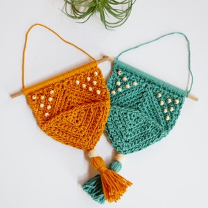 May include: Two handmade macrame wall hangings. One is mustard yellow, the other teal. Each has a wooden dowel, wooden beads, and a tassel. The macrame is a geometric pattern. The hangings are suspended by a loop of yarn.