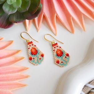 May include: A pair of beaded earrings with gold-colored hooks. The earrings feature a teardrop shape with a pattern of red, orange, and turquoise beads. The earrings are displayed on a white surface with pink and green floral accents.