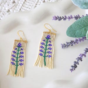 May include: Beaded dangle earrings with a floral design. The earrings feature a cream-colored base with a green stem and purple flower accents. Gold-colored hooks complete the design. The earrings are displayed on a white surface with lavender sprigs.