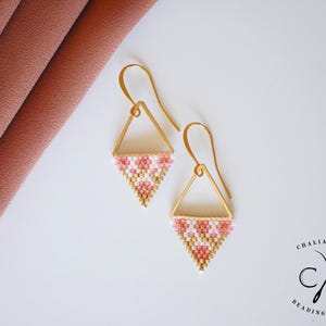 Può includere: Un paio di orecchini triangolari color oro con motivi floreali di perline rosa, bianche e oro. Gli orecchini hanno una chiusura a gancio e sono esposti su una superficie bianca.