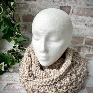 Può includere: Una sciarpa infinita di maglia beige con una trama a maglia grossa. La sciarpa è indossata intorno a una testa di manichino bianca.