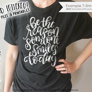 May include: A dark gray t-shirt with the text "be the reason someone smiles today" in white lettering. The text is in a handwritten style and has a distressed look.